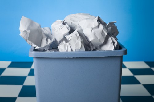 A close-up view of a light grey plastic bin filled with crumpled white paper and other waste materials, positioned on a checkered floor with a blue background. The crumpled paper shows various textures, with some pieces appearing smooth and others more wrinkled, indicates typical rubbish collected during disposal services. The bin is situated outdoors or in a warehouse environment, with the crumpled paper heaped with some pieces leaning over the edges. The scene is well-lit, highlighting the contrast between the white waste and the grey container, and subtly referencing the need for rubbish removal services such as those provided by Commercial Waste Plaistow to manage waste efficiently. The background and setting suggest a practical disposal context, aligning with general rubbish collection and waste management in the Plaistow area, postcode relevant for local service optimization.
