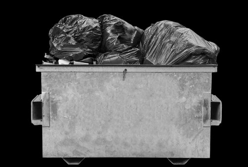 A large, sturdy waste skip situated outdoors, filled with tightly packed black plastic rubbish bags of varying sizes and materials, some with visible creases and folds. The skip, made of durable grey concrete or metal with a smooth finish, features side handles and rests on a flat paved surface, likely in a commercial or industrial area. The background is solid black, emphasizing the skip and its contents. The bags appear to contain mixed rubbish types, aligning with typical rubbish removal services, such as those offered by Commercial Waste Plaistow in East London. The lighting highlights the textures and shadows created by the bags, giving a clear view of the turbulent, crinkled surface of the plastic. This scene is representative of rubbish collection or disposal activities near a property in Plaistow, providing an accurate, detailed visual for a waste removal service context.