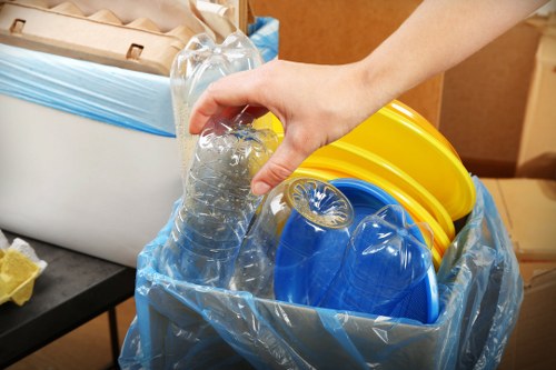A close-up image showing a person's hand holding a clear plastic bottle above a blue bin liner inside a black container, ready for disposal. The blue bin liner appears to be filled with various coloured plastic items including yellow and blue lids and containers, all made of plastic with a glossy finish. The background includes part of a white waste bin with a partially visible blue liner, an environment that appears to be indoors, possibly a storage area or warehouse. The scene is well-lit, emphasizing the textures of the plastic and the reflective surfaces of the lids. The setting suggests a rubbish collection or waste management context, highlighting the sorting of plastic waste, which is relevant to commercial waste disposal services such as those offered by Commercial Waste Plaistow in East London.