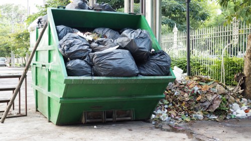 A large green industrial waste skip filled with black rubbish bags is situated outdoors on a paved surface next to a sidewalk, with some loose waste and debris scattered on the ground nearby. The skip's metal surface shows signs of use and weathering, and it is positioned close to a metal fence and some green foliage, including trees and bushes, indicating a residential or urban area in Plaistow. In the background, a white railing and additional trees can be seen, suggesting a park or communal space nearby. The scene is lit by natural daylight, highlighting the contrast between the dark rubbish bags and the vibrant green of the skip, reflecting a typical rubbish collection setting managed by a commercial waste service like Commercial Waste Plaistow, serving local businesses and residents in the postcode area.