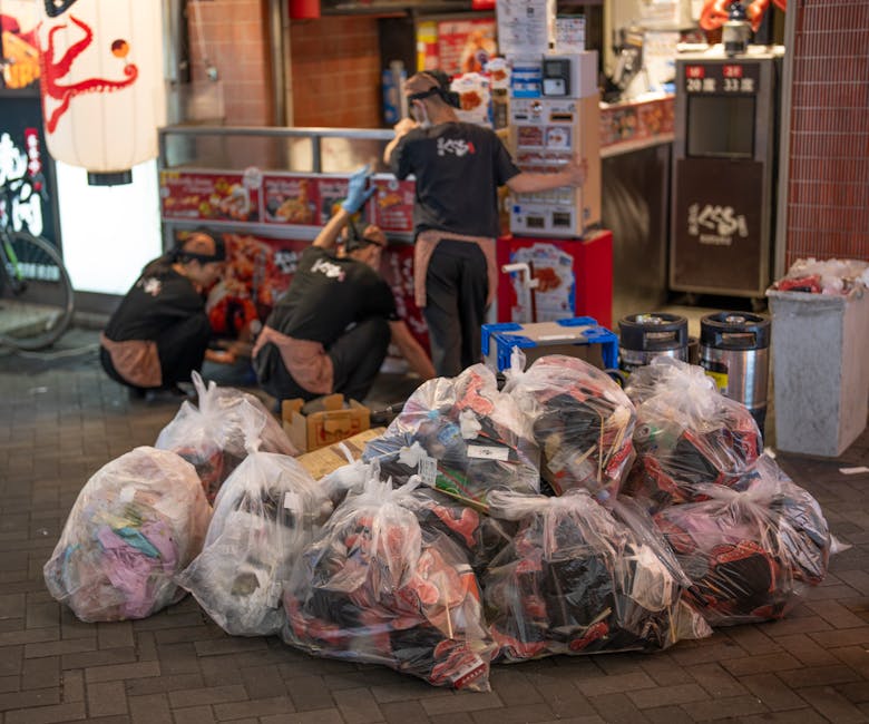 A stack of various discarded waste materials placed on a concrete driveway in an outdoor setting. The pile includes large black garbage bags made of glossy plastic, filled with refuse, and some are ti