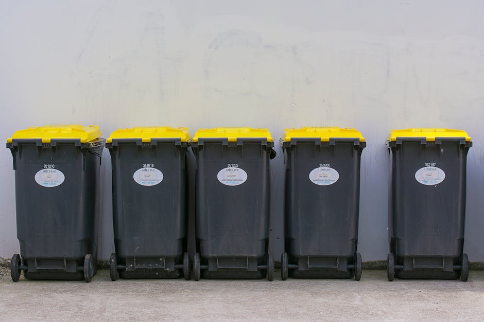A middle-aged man with dark hair, wearing a black T-shirt with white text on the front, is seen disposing of waste into a tall, stainless steel trash bin located on a paved outdoor walkway. He is hold